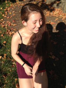 A photo of Erin smiling from above. She wears her hair down and black camisole and shorts. Under her is grass and leaves.