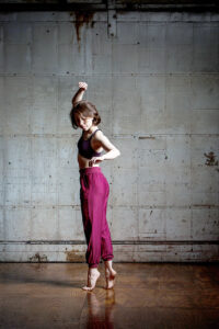 Erin in a dance studio on releve with one arm lifted. She wears a black tank top and baggy purple pants.