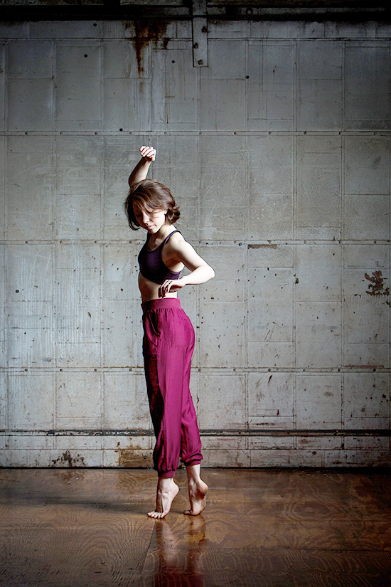 Erin in a dance studio on releve with one arm lifted. She wears a sports bra and baggy purple pants.