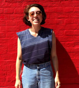 Erin smiles at the camera standing against a red brick wall. She wears sunglasses, a tank top, and jeans.