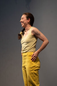 A dancer wearing yellow pants and tank top stands and smiles to the side. Her hair is in a braid and her hands are on her hips. The background is gray.