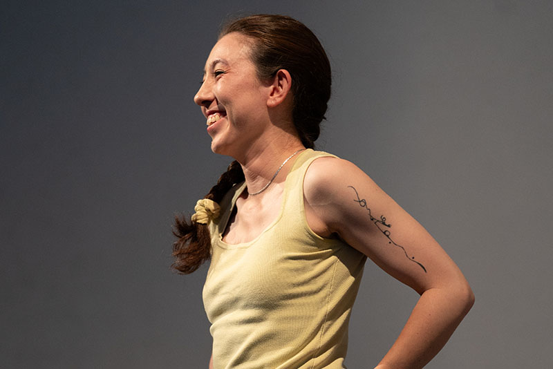 A dancer wearing yellow pants and tank top stands and smiles to the side. Her hair is in a braid and her hands are on her hips. The background is gray.