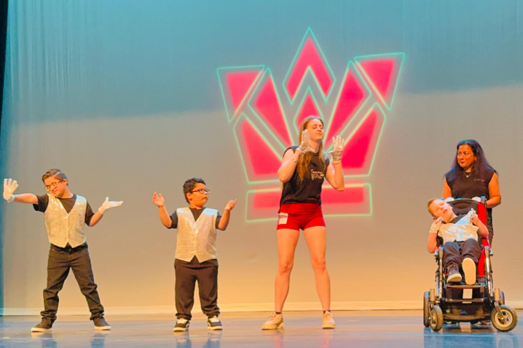 Five performers, including two children in matching silver vests and an individual using a wheelchair, pose together on stage in front of a brightly lit crown backdrop.