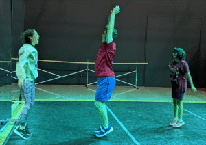 Three people leap simultaneously in a gym-like room with a ballet barre, their bodies captured mid-air in an energetic, synchronized jump.