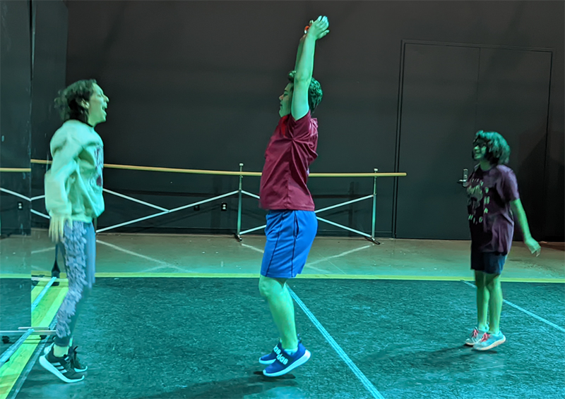 Three people leap simultaneously in a gym-like room with a ballet barre, their bodies captured mid-air in an energetic, synchronized jump.