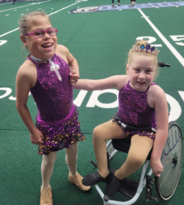 Two girls in sparkly purple costumes pose on a football field. One is in a wheelchair, and the other is standing next to her.
