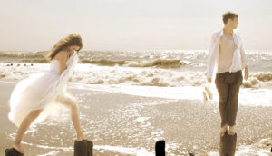Two individuals stand on wooden posts at the beach with waves crashing behind them; one wears a white dress, and the other is in casual attire holding shoes.