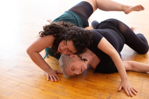 Two dancers roll together on a wooden floor, their heads connected.