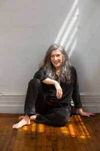 A person with long hair sits on a wooden floor against a wall, one leg bent and the other extended, dressed in black clothing.