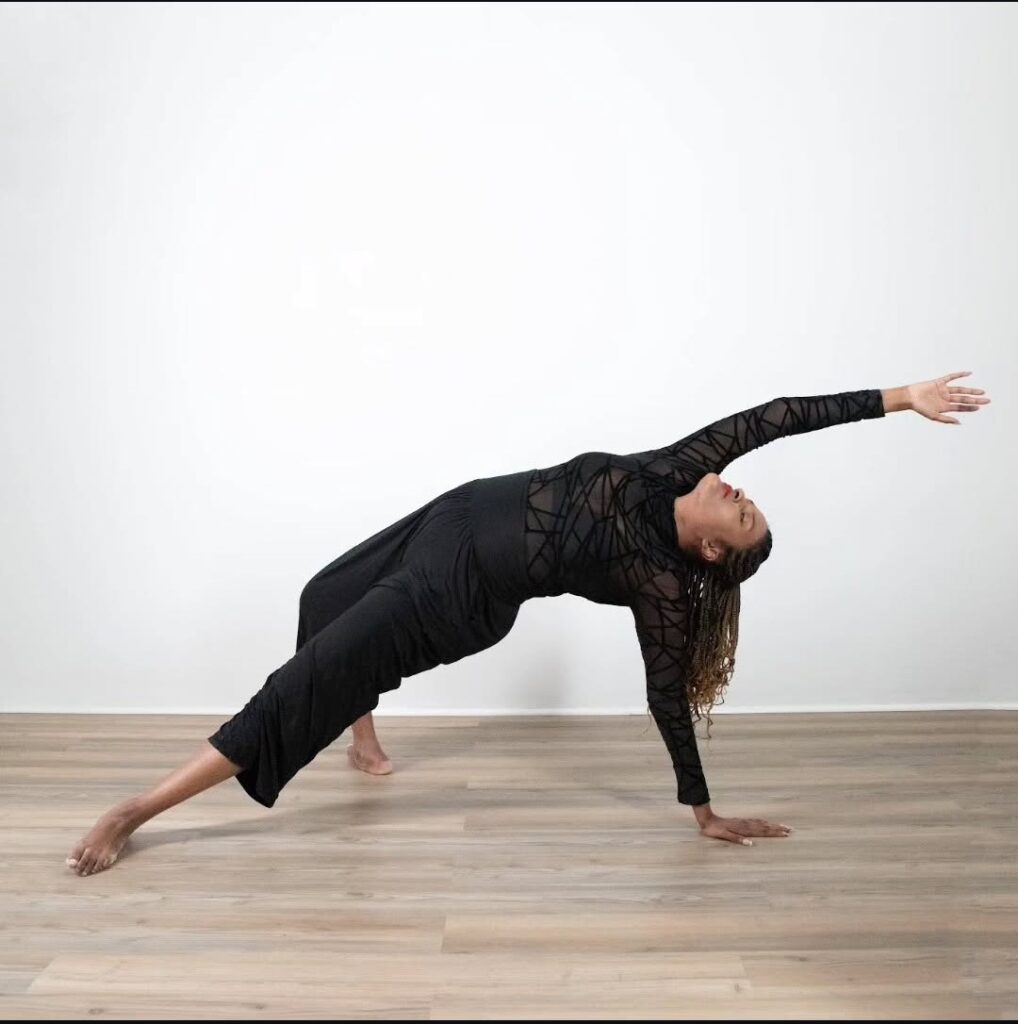 Devin leans back and supports their weight on one hand and one foot, their other hand and foot extended outward. They are wearing black in a dance studio.