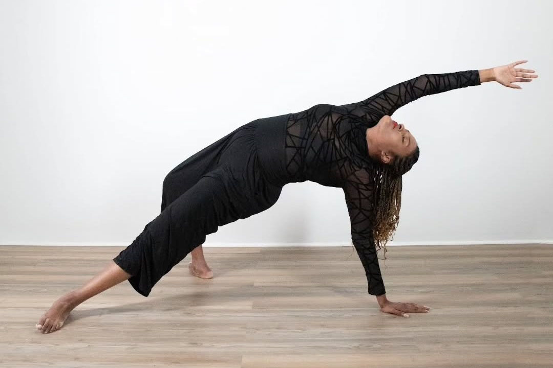 Devin supports their weight on one hand and one foot, their other hand and foot extended outward. They are wearing black in a dance studio.