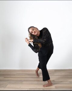 Devin lunges forward and leans to the side, their hands close together to the side. They are wearing black in a dance studio.