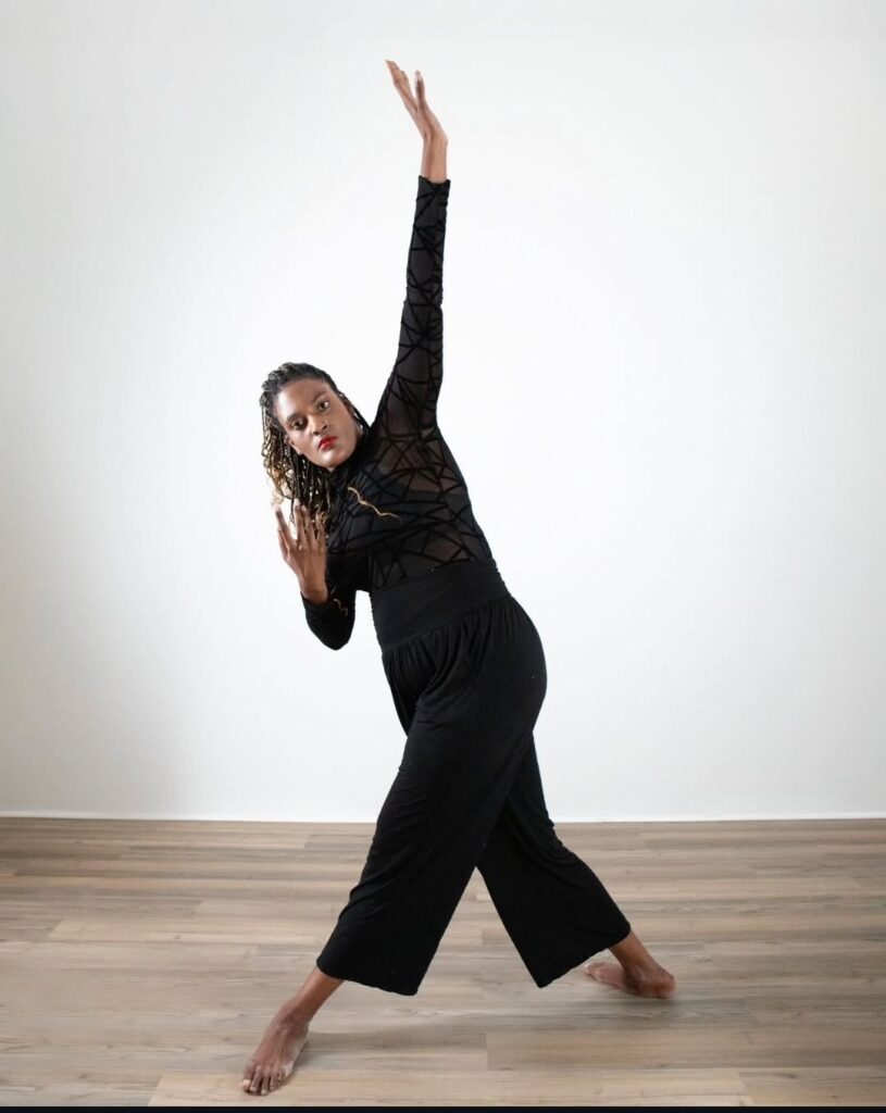 Devin stands with legs far apart on the diagonal, one arm raised and the other bent by their ear. They are wearing black in a dance studio.