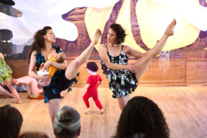 Two dancers onstage lift their legs to the side. One is holding an infant. A toddler runs in the background.