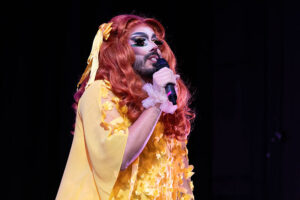 Eric Garcia onstage dressed in drag makeup and wearing a yellow dress, holding a microphone.