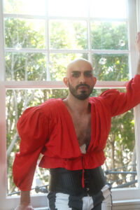 Eric Garcia wears a red billowing blouse and black and white pants. He stands in front of a window with greenery.