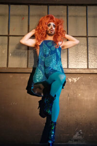 Eric Garcia onstage wearing a turquoise dress and leggings, drag makeup, and a red wig.