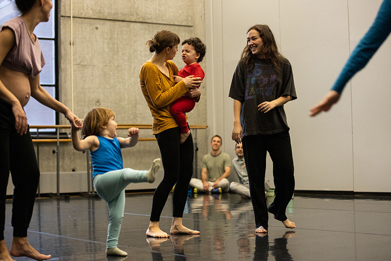 Three adult dancers, one of whom is pregnant, stand and walk around two toddlers, one of whom is being held and the other is doing a big kick.