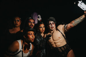 Six performers wearing cabaret costumes peer forward out of the darkness at a light one of the performers is holding.