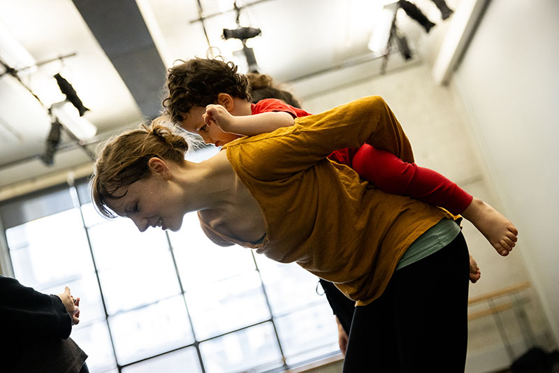 Esme is in a dance studio giving her toddler son a piggy back ride.