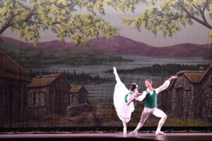 Sydney and her partner do a penchee arabesque onstage against a backdrop that looks like a village.