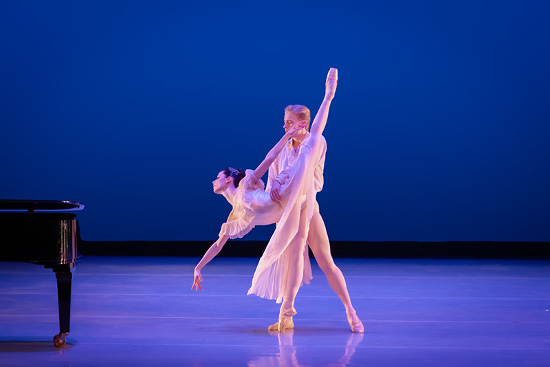 Sydney is in a penche arabesque onstage, supported by her partner at the waist. They are both wearing flowy white costumes. A piano is in the corner.