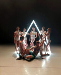 Seven dancers onstage in front of a glowing white triangle.