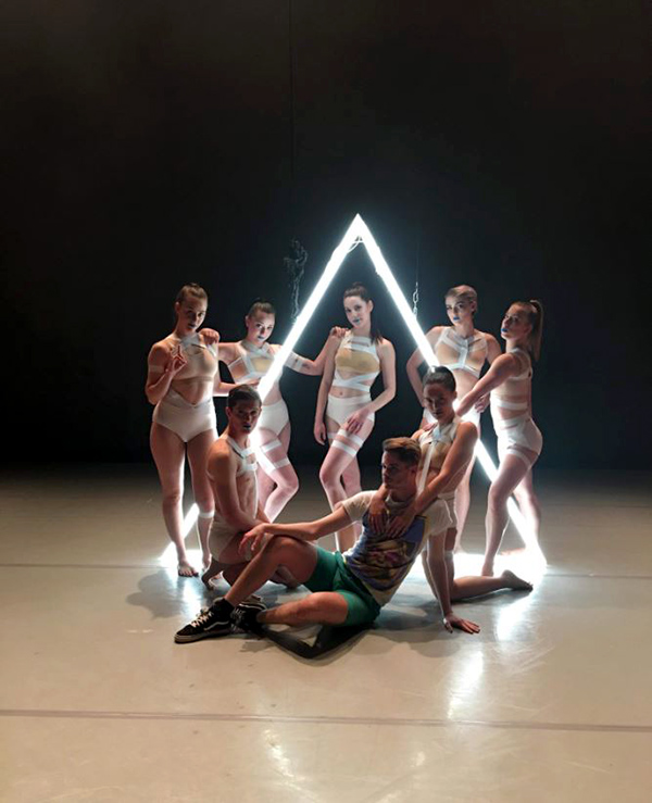 Seven female dancers in white costumes and one man sitting on the floor in shorts and t-shirt onstage in front of a glowing white triangle.