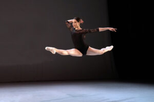 Sydney jumps through the air in a split onstage wearing a black leotard and pink tights.