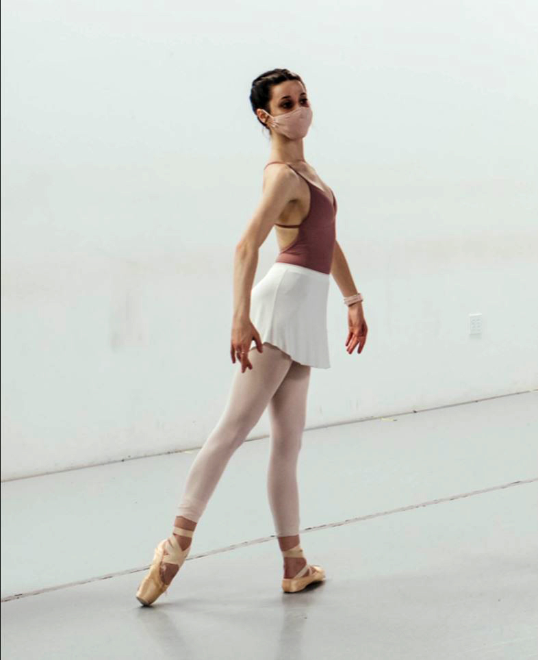 A dancer in tendu arabesque in a studio wearing a mask.
