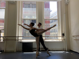 Sydney is partnered in an arabesque by another dancer in front of a window in a dance studio.