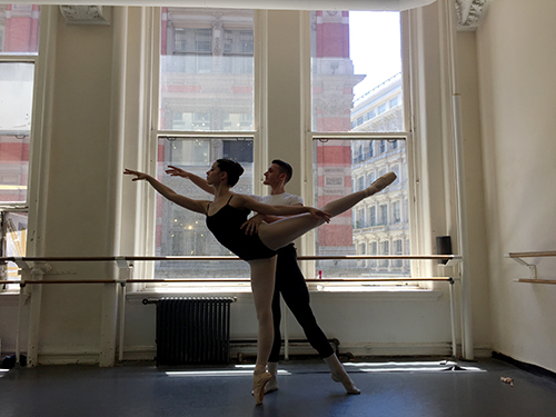 Sydney is partnered in an arabesque by another dancer in front of a window in a dance studio.