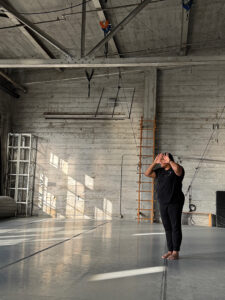 Bhumi stands in a big dance studio wearing black. Her hands are in front of her face and she peers though her fingers.