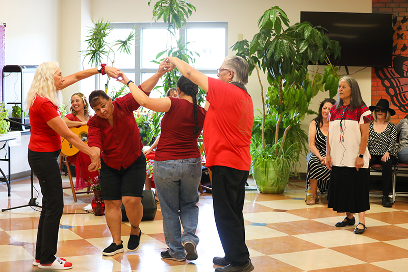 Lucy stands to the side and coaches four dancers looping their arms around each other. A guitarist plays in the background.