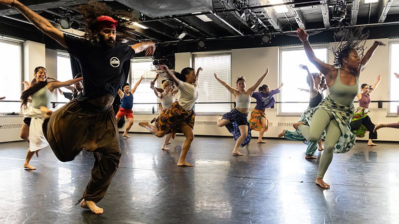 Several dancers in a studio move across the floor with arms extended to the side and one foot behind them.