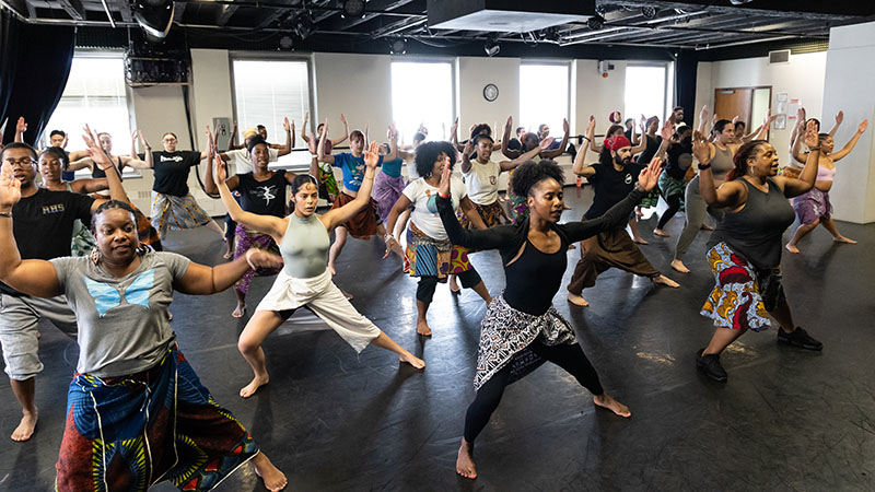 Several dancers in a studio lunge to one side and lift their arms to their sides.