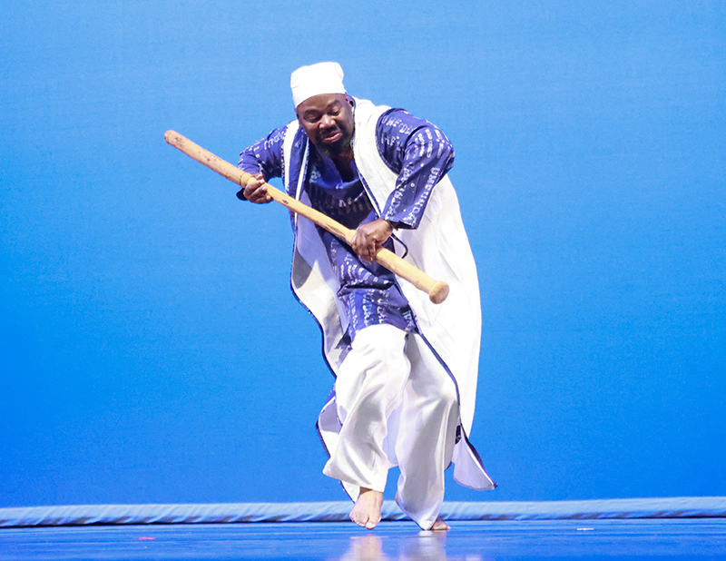 A dancer onstage wearing white and purple hunches forward holding a long stick and walking forward.