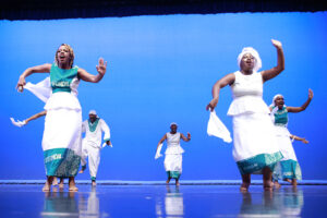 Two dancers onstage in white with blue accents lift one arm and hold a white kerchief in the other arm. Three dancers mirror them in the background.
