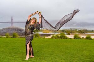 A belly dancer stands on grass with her arms over her head holding a long sheer scarf trailing in the wind. The Golden Gate bridge is in the background.