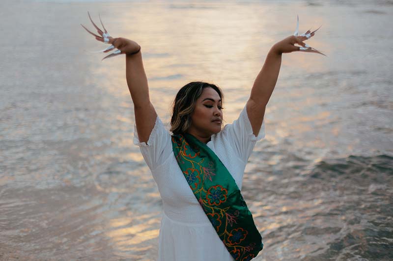 A dancer from the waist up wearing a white shirt and a green sash. Her arms are extended above her head and her hands flair out with long fingernails. An ocean is behind her.