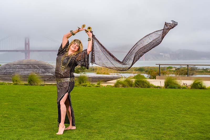 A belly dancer stands on grass with her arms over her head holding a long sheer scarf trailing in the wind. The Golden Gate bridge is in the background.