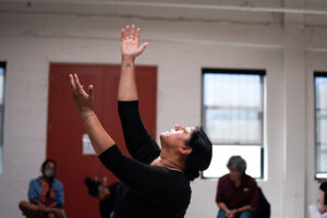 Bhumi looks up and extends to arms upward. She is in a dance studio. People sit and watch in the background.