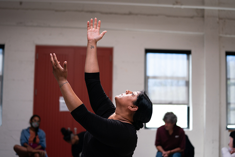 Bhumi looks up and extends to arms upward. She is in a dance studio. People sit and watch in the background.