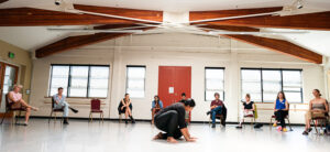 A dancer wearing black kneels with hands to the floor in a big dance studio. People sit and watch in a line of chairs in the background.