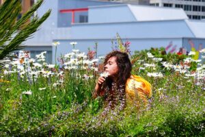 A dancer with long brown hair and wearing yellow crouches down in a field of flowers and lifts a white flower to their nose.