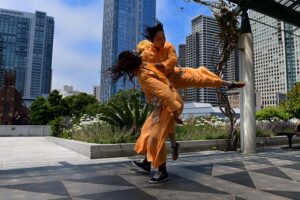 One dancers lights another dancer who shoots their leg out to the side. They are wearing orange and a city scape rises behind them.