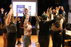 A group of people stand in a circle with their arms overhead. They are in a big room with a wood floor and a projector in the background.