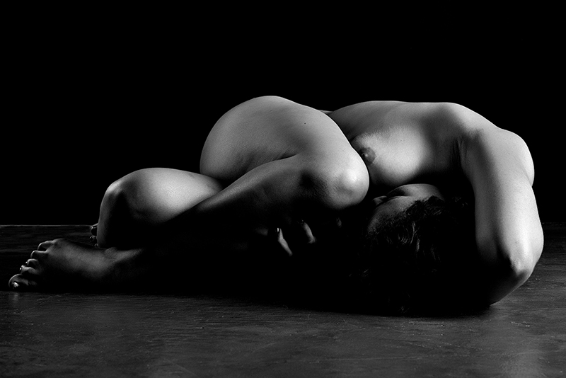 A person lies naked on a floor curled up with one knee to their forehead and one arm over their hair.