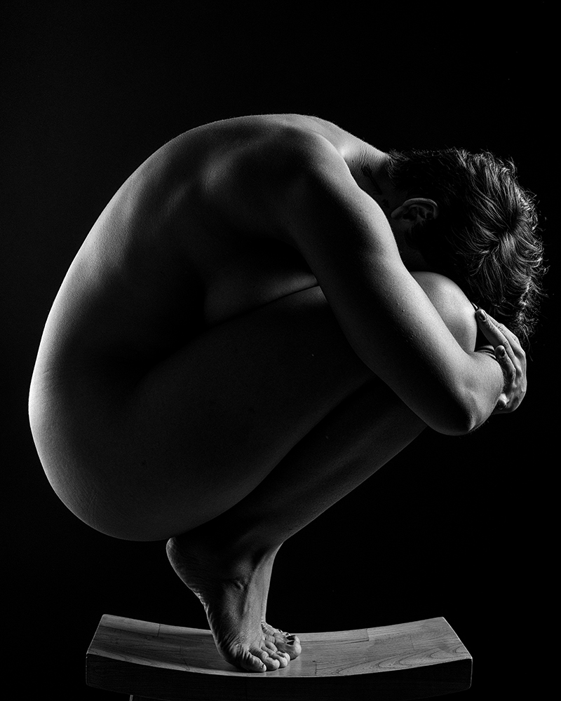 A profile of a a naked body on tip toes hunched over and embracing their knees.