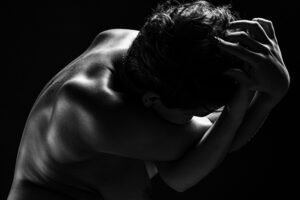 A naked back and head in low light, the arms twisted around each other toward the head.